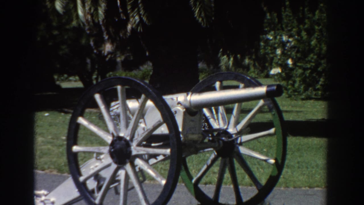 Silver cannon with large spoked wheels on a path beside grass and trees.