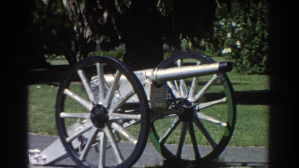 The Perth park cannon remains centered in a grainy 8mm frame, with lawn and foliage behind it.