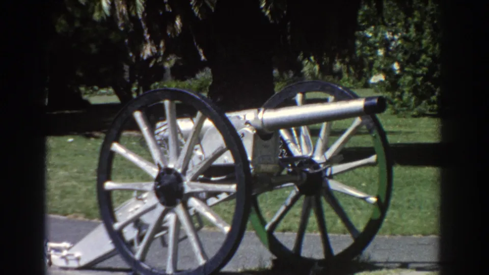 Final 8mm frame of the Perth park cannon display, with the silver barrel, spoked wheels and shaded foliage visible.