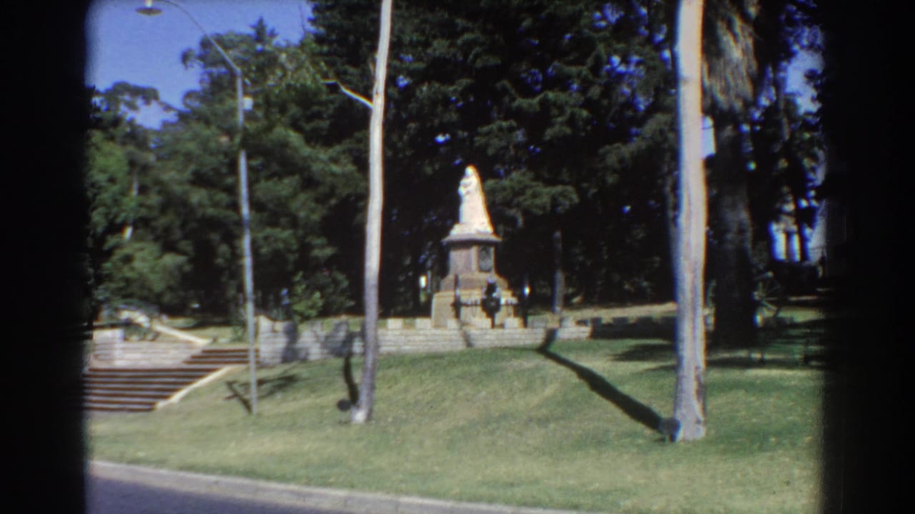 1967 Perth home-movie frame showing a distant park statue on a stone pedestal, bordered by steps, lawn, trees, and a lamppost.