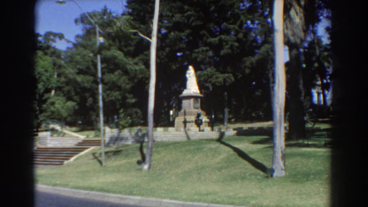 Park monument seen across grass, with stone steps at left and tall trees behind.