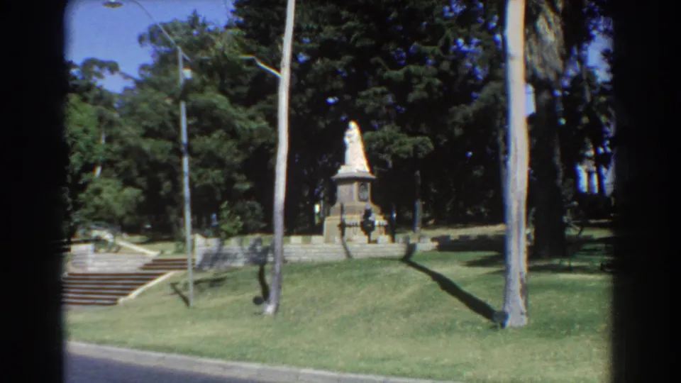 1967 Perth 8mm frame of a statue-topped monument across a sunlit lawn, with steps, a lamppost, and dark evergreens behind it.