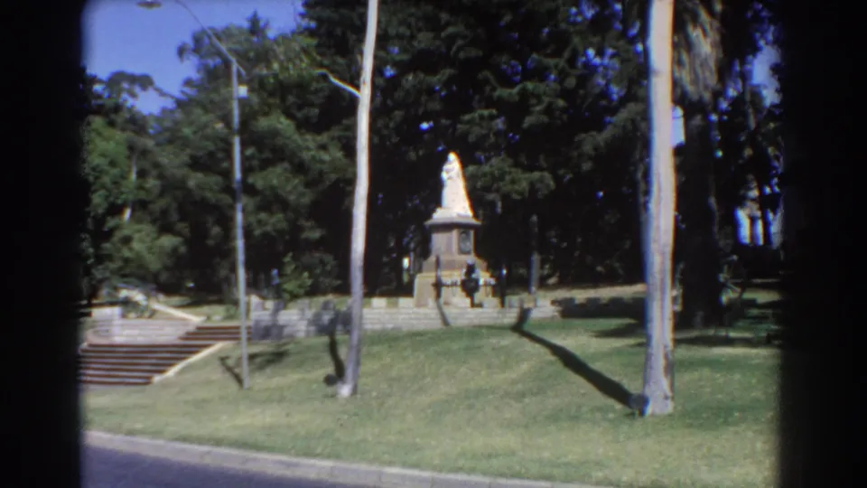Wide 1967 Perth home-movie view of the park memorial from below, showing the statue, pedestal, steps, lawn, lamppost, and tall trees.