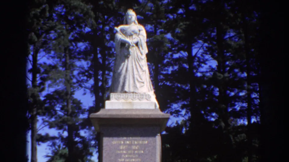 Closer 1967 Perth frame showing a pale robed statue on a stone pedestal, backed by dense trees and blue sky.