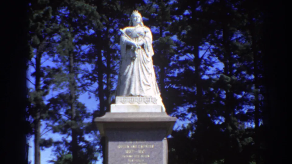 1967 Perth 8mm frame showing the robed statue, pedestal face, and surrounding pine-like trees in soft focus.