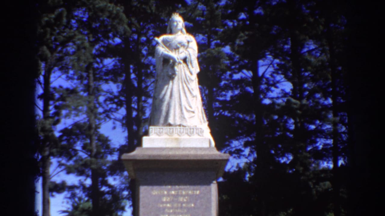 Queen Victoria statue on a stone pedestal among tall trees under blue sky.