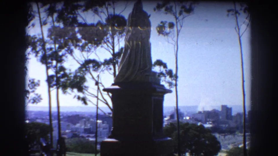 1967 Perth 8mm frame showing a dark statue and pedestal against trees, blue sky, and distant city buildings.