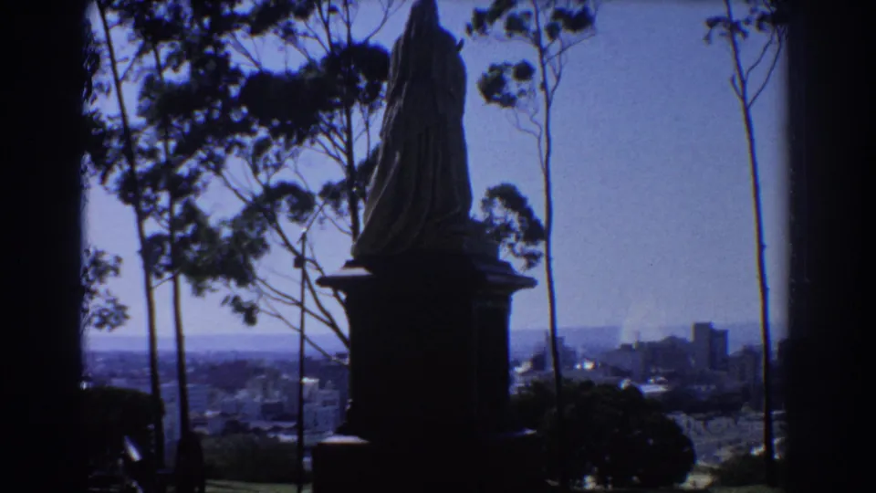 1967 Perth 8mm frame holding on the statue silhouette, with trees and a hazy cityscape beyond.