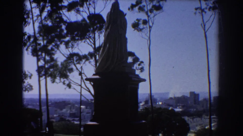 1967 Perth overlook with a silhouetted statue, narrow tree trunks, and distant urban buildings.