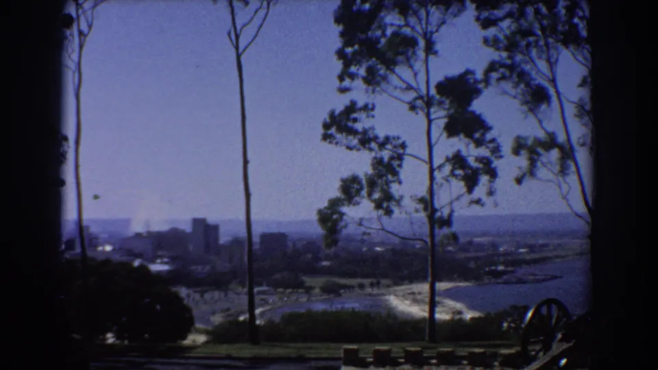 1967 Perth home-movie frame looking across water and shoreline toward distant buildings, with tall trees in the foreground.