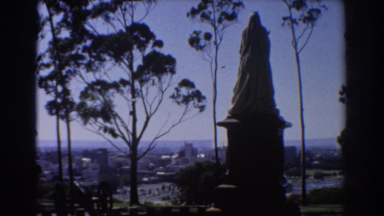Silhouetted statue on a plinth beside eucalyptus trees with a distant city skyline below.
