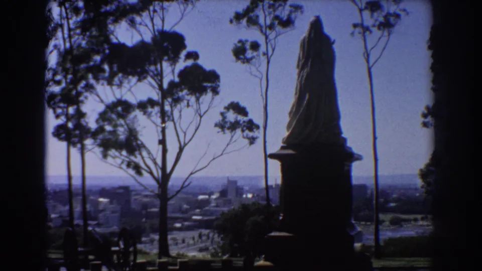 Hilltop Perth view in 1967 with a statue in silhouette, eucalyptus trunks, and the city spread across the horizon.
