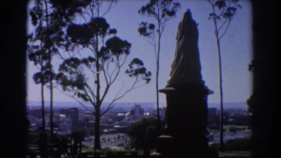 1967 Perth lookout scene showing a robed statue in silhouette, tall eucalyptus trees, and a soft distant cityscape.