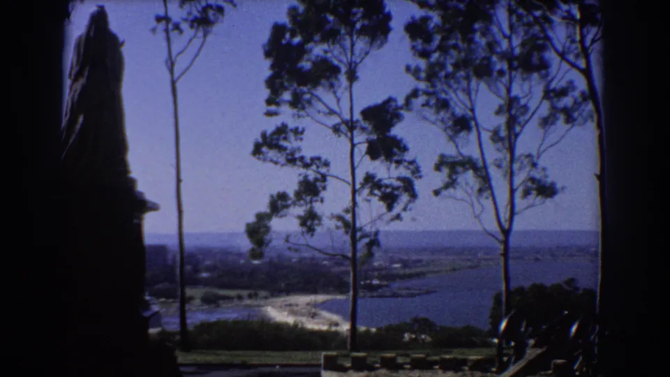 1967 Perth 8mm frame showing a coastal panorama through tall trees, with old cannons in the shaded foreground.
