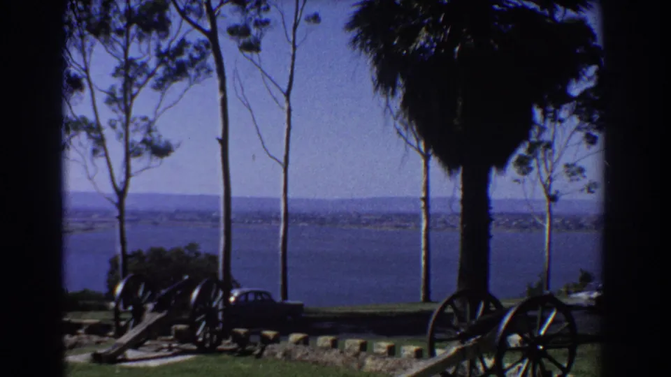 1967 Perth home-movie view across water from a park-like overlook with trees, cannons, grass, and a parked car.