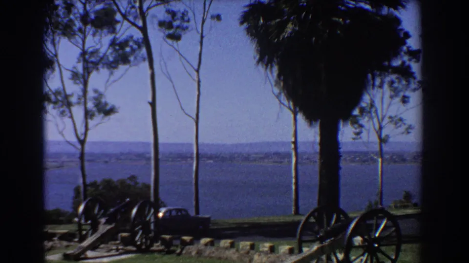 1967 Perth 8mm frame showing trees, cannons, a partial parked sedan, and a broad waterfront under clear sky.