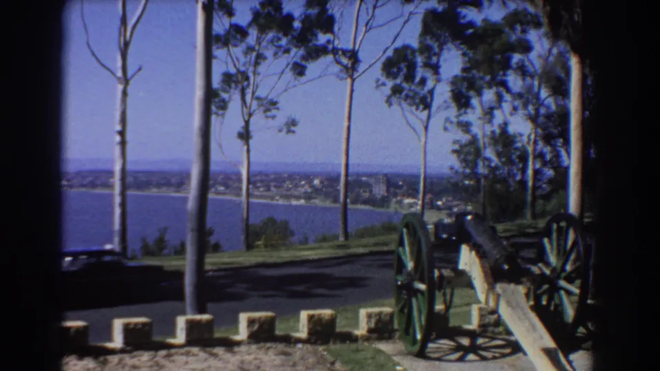 Black classic car passes near a cannon at a Perth overlook, with lake water and distant buildings beyond the trees.