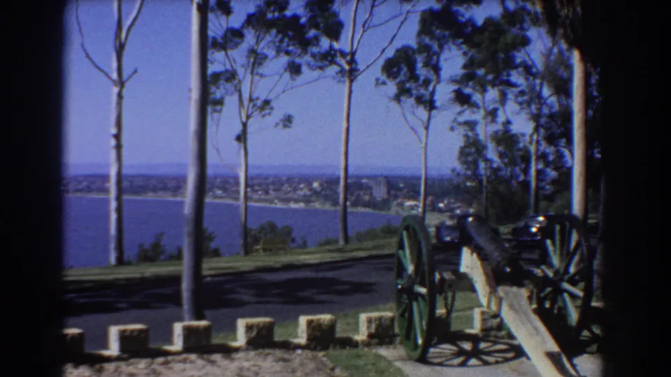 Perth 1967 home-movie frame of a wheeled cannon at a sunny overlook, with trees, water, and shoreline buildings behind it.
