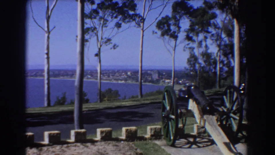 1967 Perth 8mm frame showing a cannon with green wheels in a park above the sea and distant shoreline.