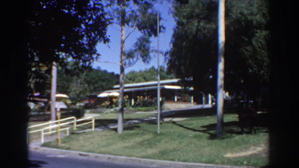 1967 Perth 8mm view of a landscaped park area with railings, trees, a low building, and small distant visitors.