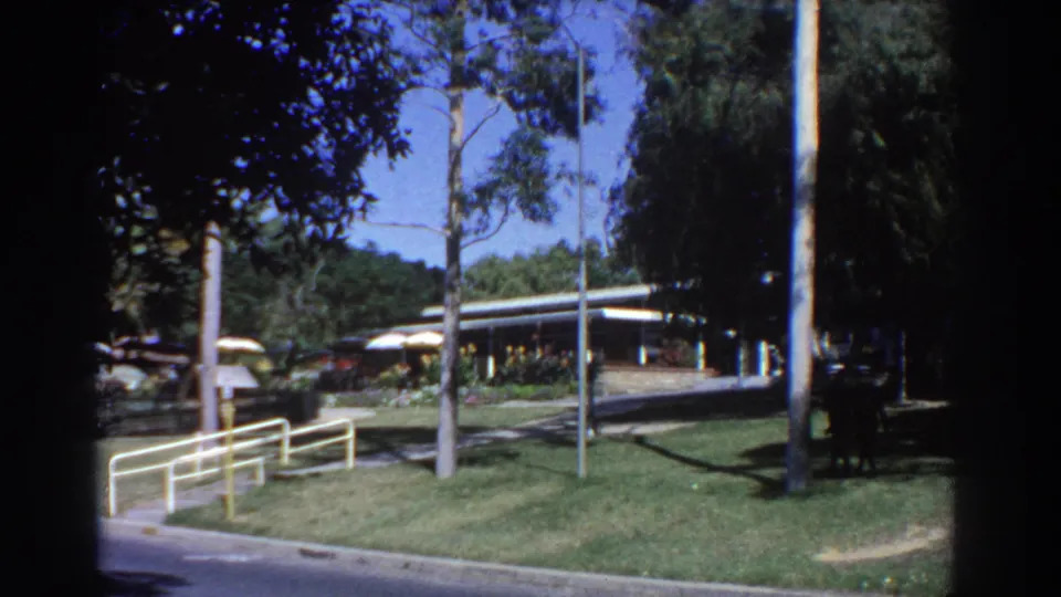 1967 Perth park frame showing a road curve, lawn, railings, eucalyptus trees, low building, and distant people.