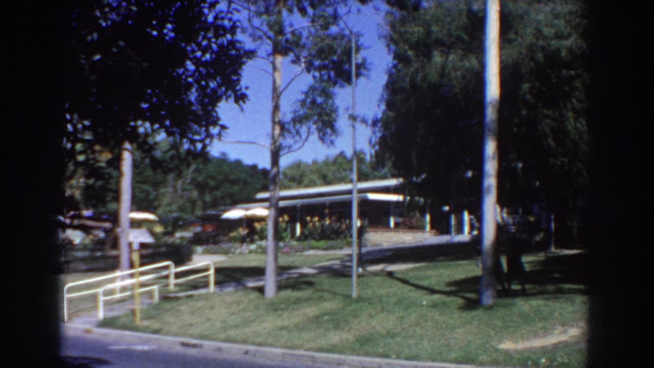 Modern flat-roof building behind lawns, white railings, trees, and a distant pedestrian.