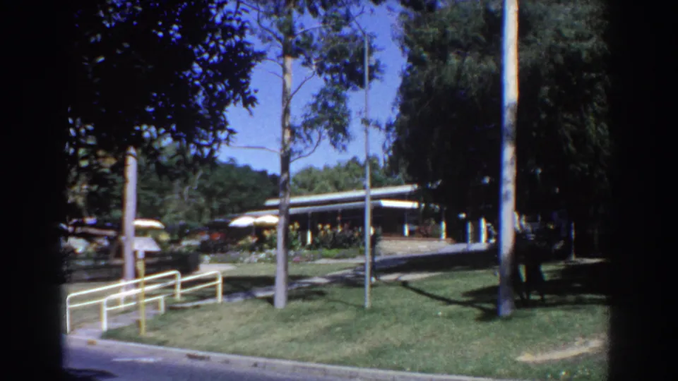 1967 Perth home-movie frame showing a modern low building, outdoor umbrellas, lawn, railings, trees, and a person walking nearby.