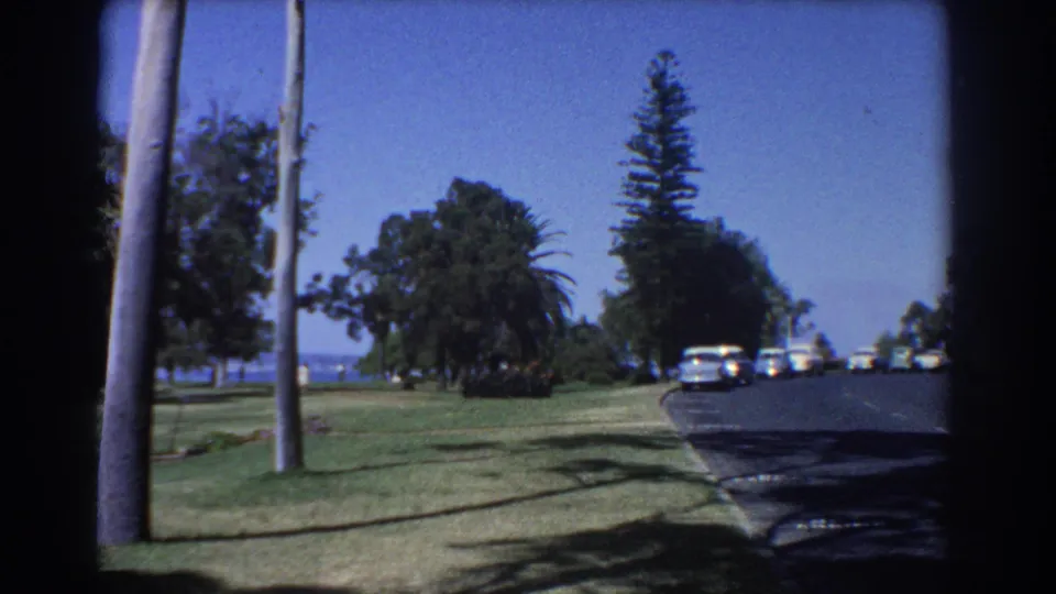 Late-1960s Perth home-movie frame showing a coastal road, roadside lawns, palms, tall trees, distant water, and period cars.
