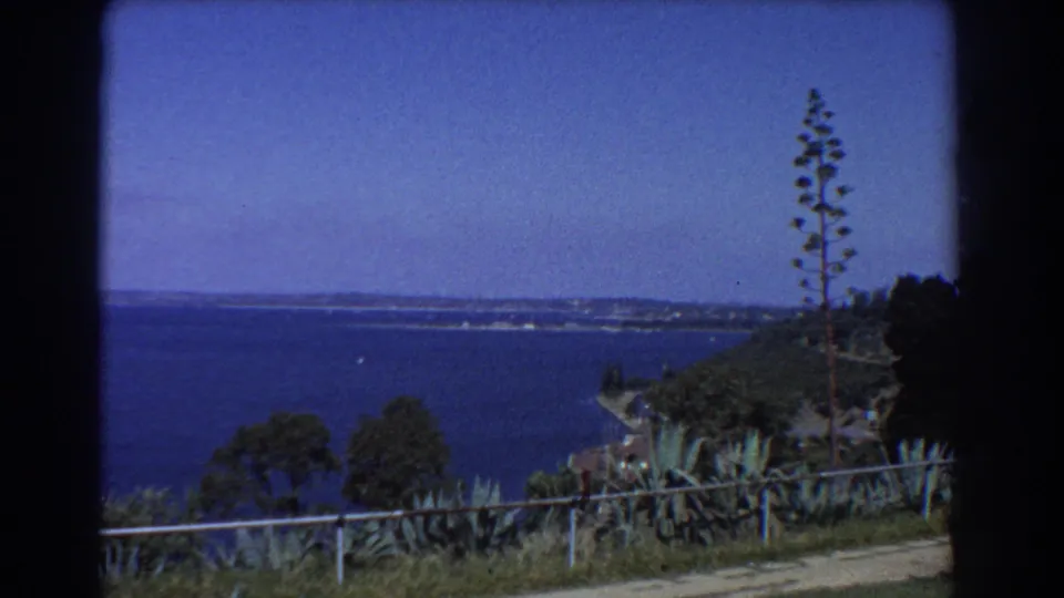 1967 Perth sea view over a low fence, with coastal vegetation, a path, and distant shoreline under clear sky.