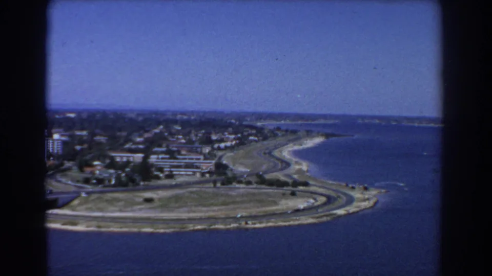 Aerial view over Perth, Australia showing a curved shoreline, waterfront roads, open land, beach edge, and low urban buildings.