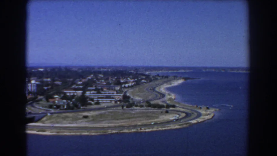 1967 Perth aerial frame showing blue water, a sweeping road along the shoreline, beach margin, open ground, and nearby residential development.