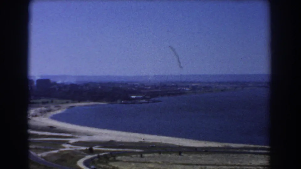 1967 8mm coastal view near Perth with a curved beach, broad blue water, low shoreline and open land below.