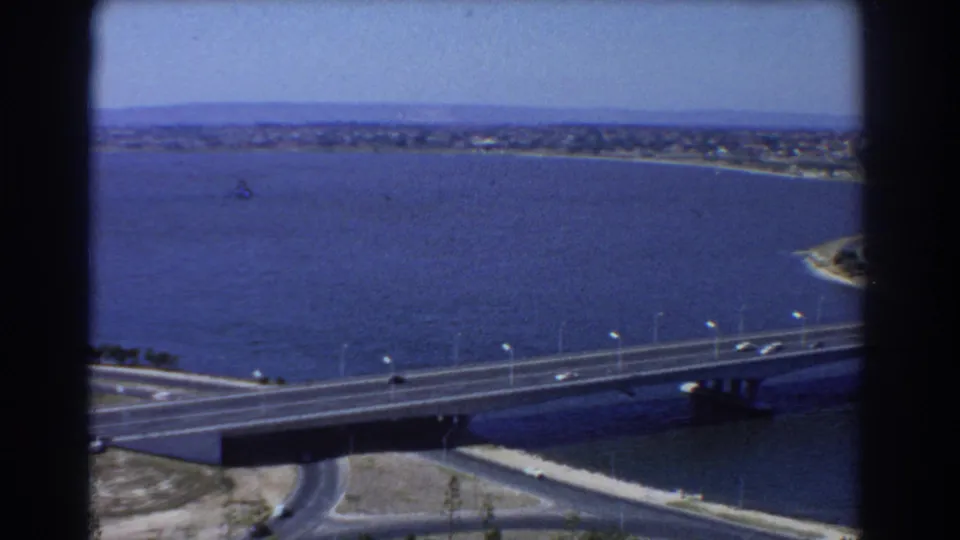 1967 Perth aerial frame with a long road bridge over a broad waterway, light vehicle traffic, shoreline, and distant buildings.