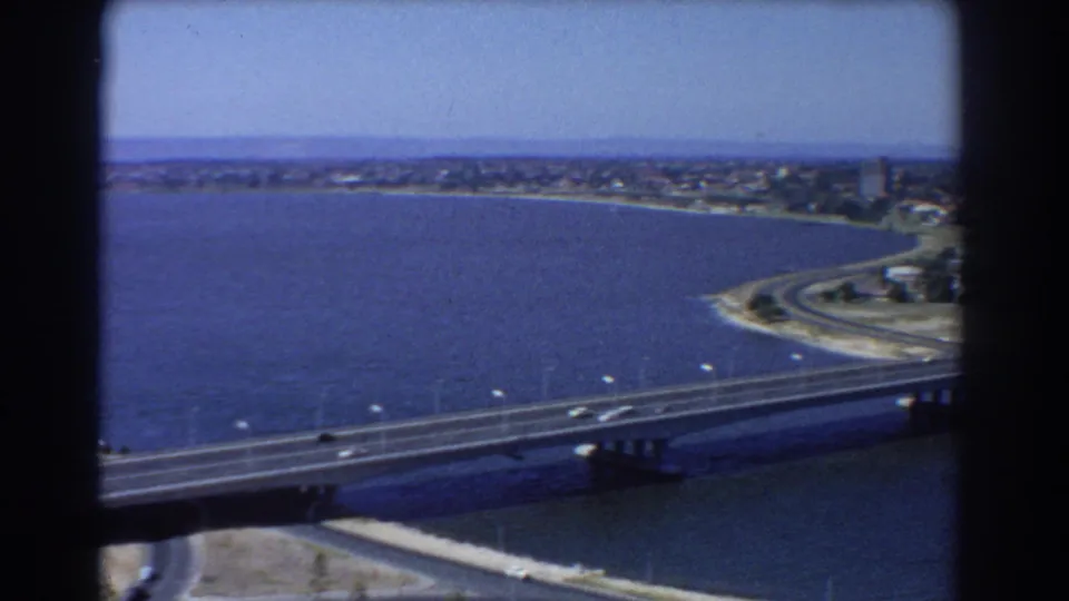 Aerial 8mm view of Perth waterfront with bridge lanes, blue water, a curving shore road, and distant low-rise development.