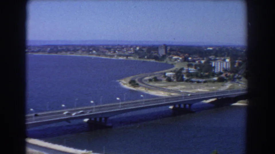 1967 Perth aerial frame showing the bridge foreground, waterfront roads, open water, and distant coastal buildings under clear sky.