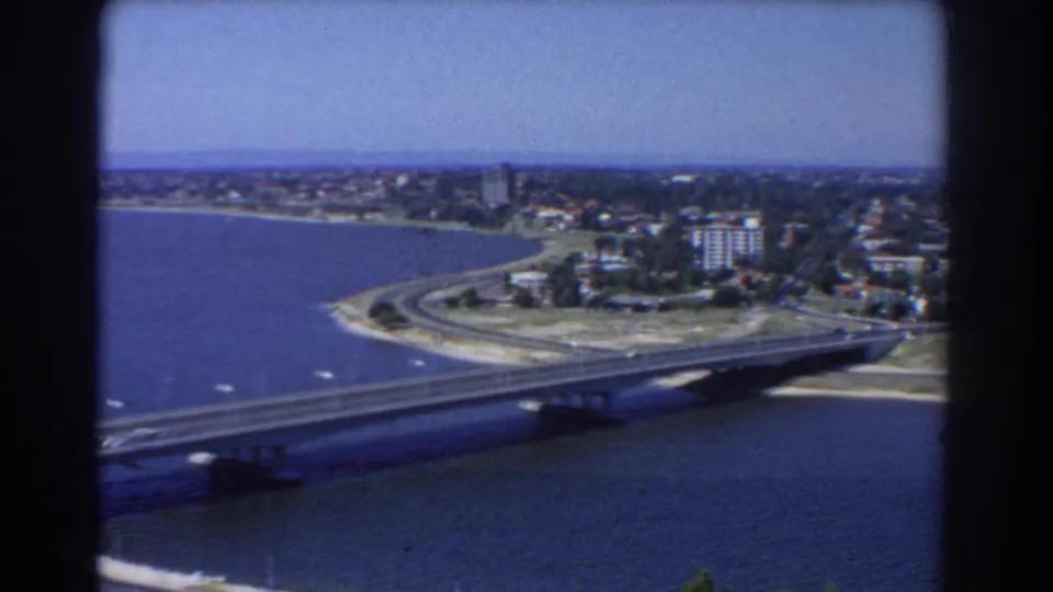 1967 Perth view from above, with a bridge over blue water, shoreline roads, low-rise buildings, and distant suburbs.