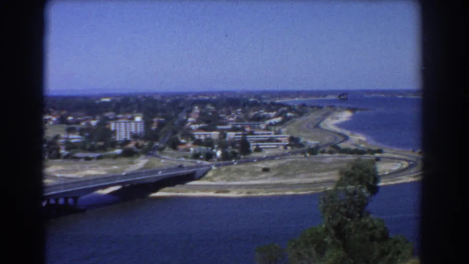 Elevated 1967 Perth view showing the edge of a bridge, a curving road network, shoreline development, blue water, and distant land.
