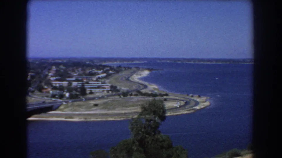 1967 Perth shoreline view with blue water, a curving road, low-rise buildings, distant shoreline, and leafy foreground partially covering the frame.