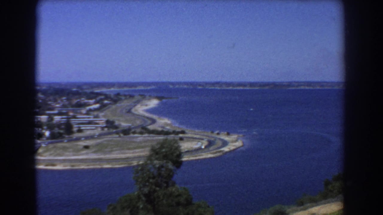 1967 Perth home-movie frame looking over blue ocean water, pale sand, a curved coastal road, and distant residential shoreline.