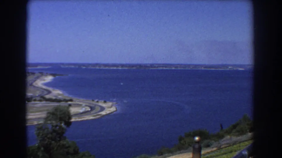 1967 Perth coastal panorama showing a sandy point, winding shoreline road, deep blue water, hillside vegetation, and distant horizon.