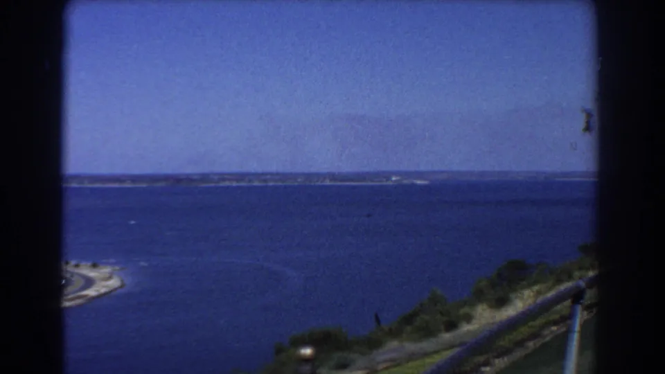 1967 Perth lookout view across blue water toward a distant shoreline, with hillside vegetation and a metal railing in the foreground.