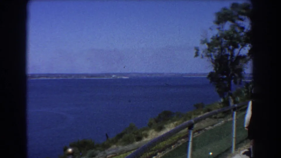1967 Perth home-movie frame showing a hillside lookout over blue coastal water, with railing, trees, distant land, and clear sky.