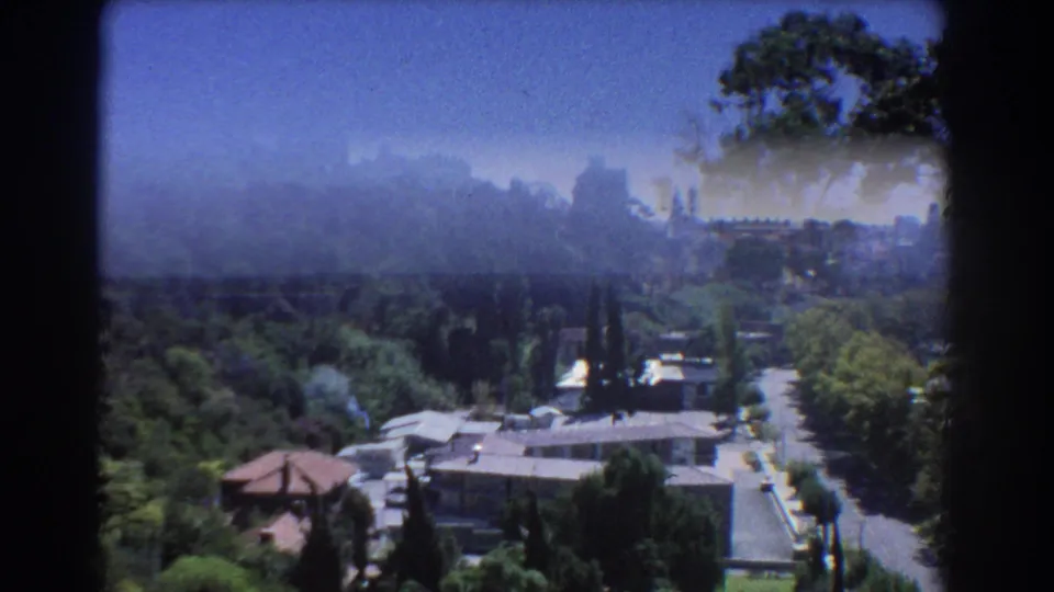 1967 Perth home-movie frame looking across a wooded hillside, low buildings, road, and distant high-rises under haze.
