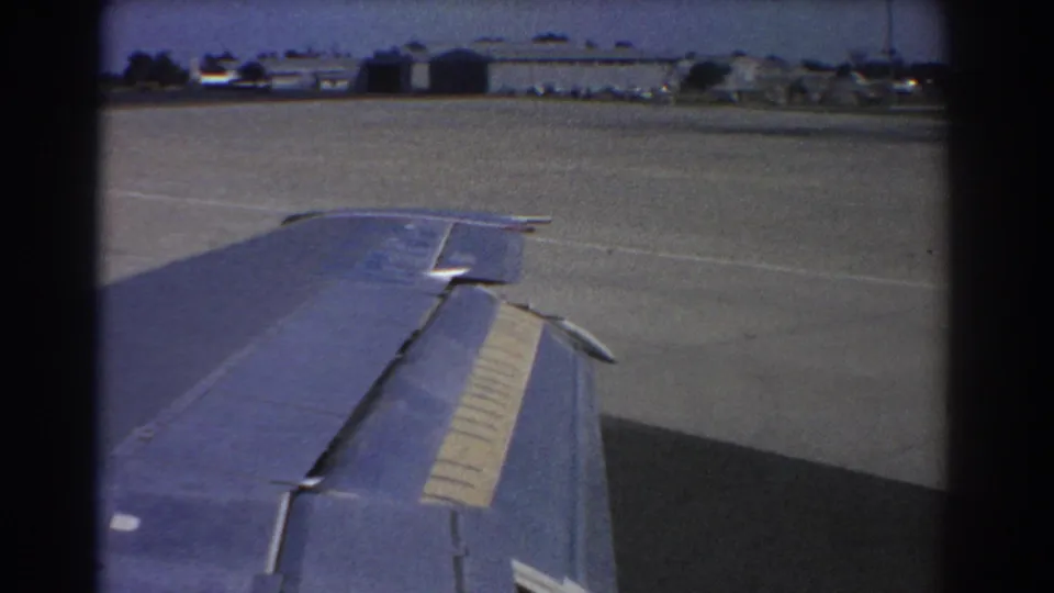 1967 Perth home-movie frame showing a blue aircraft wing, paved tarmac, and low airport buildings in the distance.