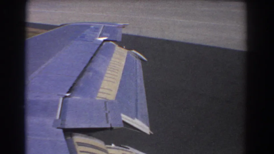 Aircraft window view in the 1967 Perth clip, with the blue wing and flap surfaces filling most of the frame over runway pavement.