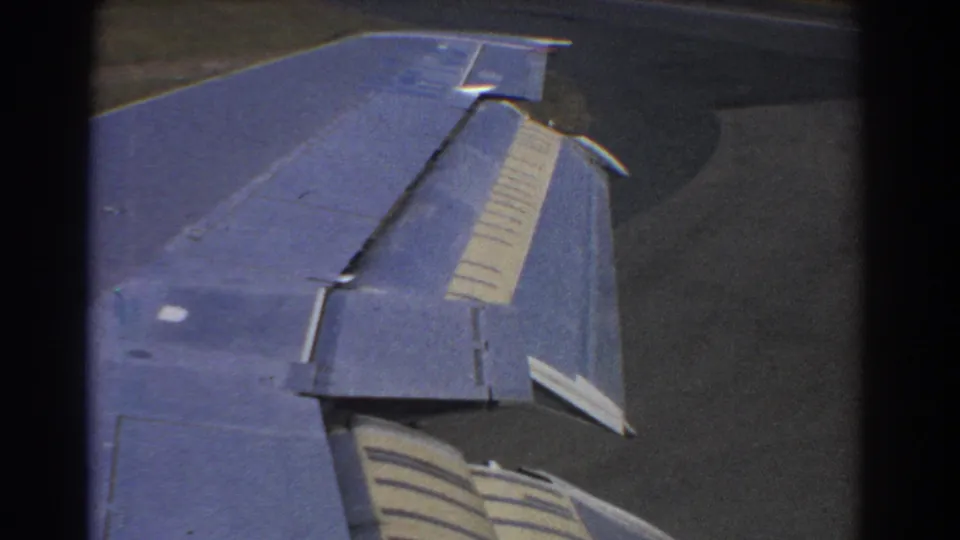 Final 1967 Perth window view of the blue aircraft wing and flap over dark tarmac, with visible 8mm frame edges.