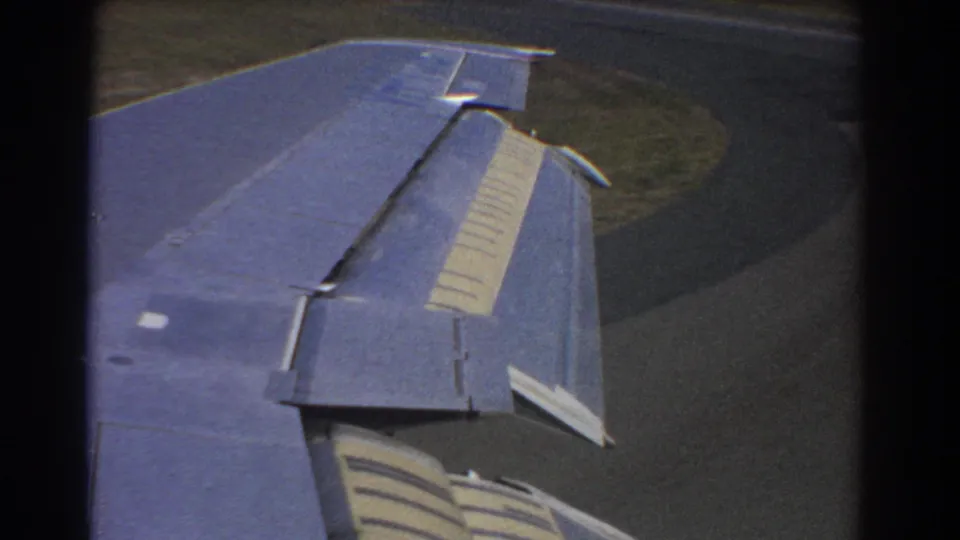 1967 Perth, Australia 8mm frame showing a blue aircraft wing and striped flaps above runway and grassy ground.