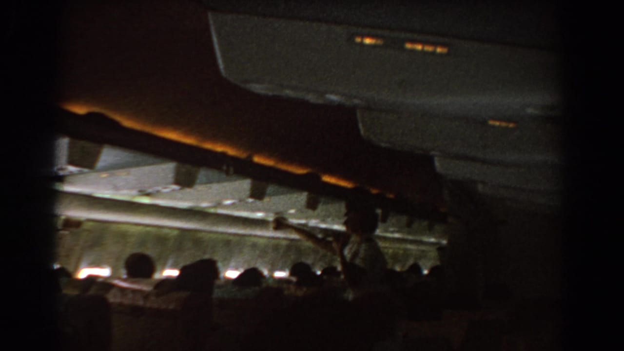 Dim airplane cabin with seated passengers and a flight attendant near the aisle.