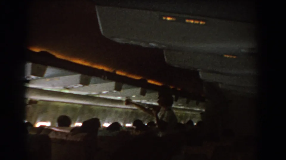1967 home-movie view inside a commercial airliner cabin, with passengers seated as a flight attendant gestures near the overhead area.