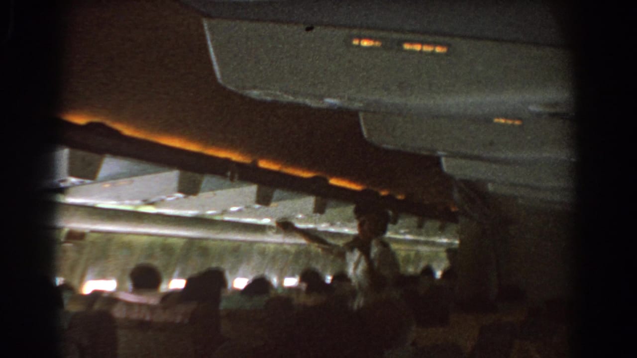 Flight attendant points upward in a dim aircraft cabin as passengers sit below.